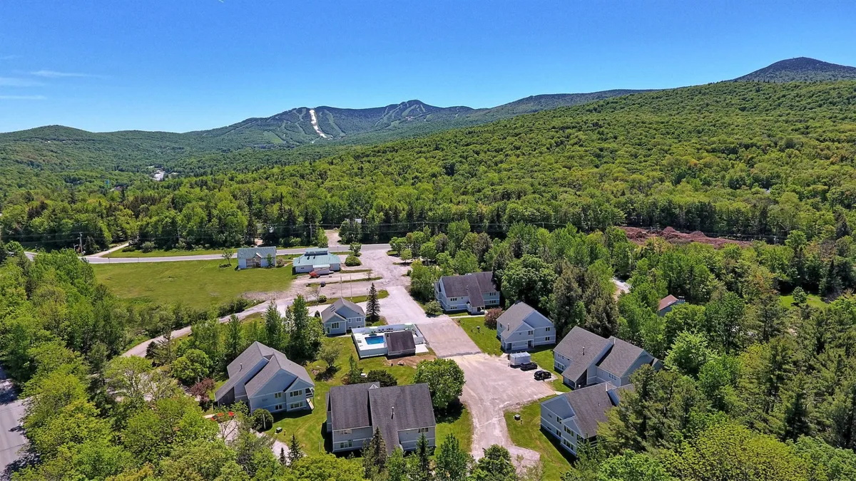 This exterior image captures a serene Vermont landscape with green mountains in the background, a cluster of homes nestled among lush foliage, and open space that hints at nature trails and outdoor activity possibilities.