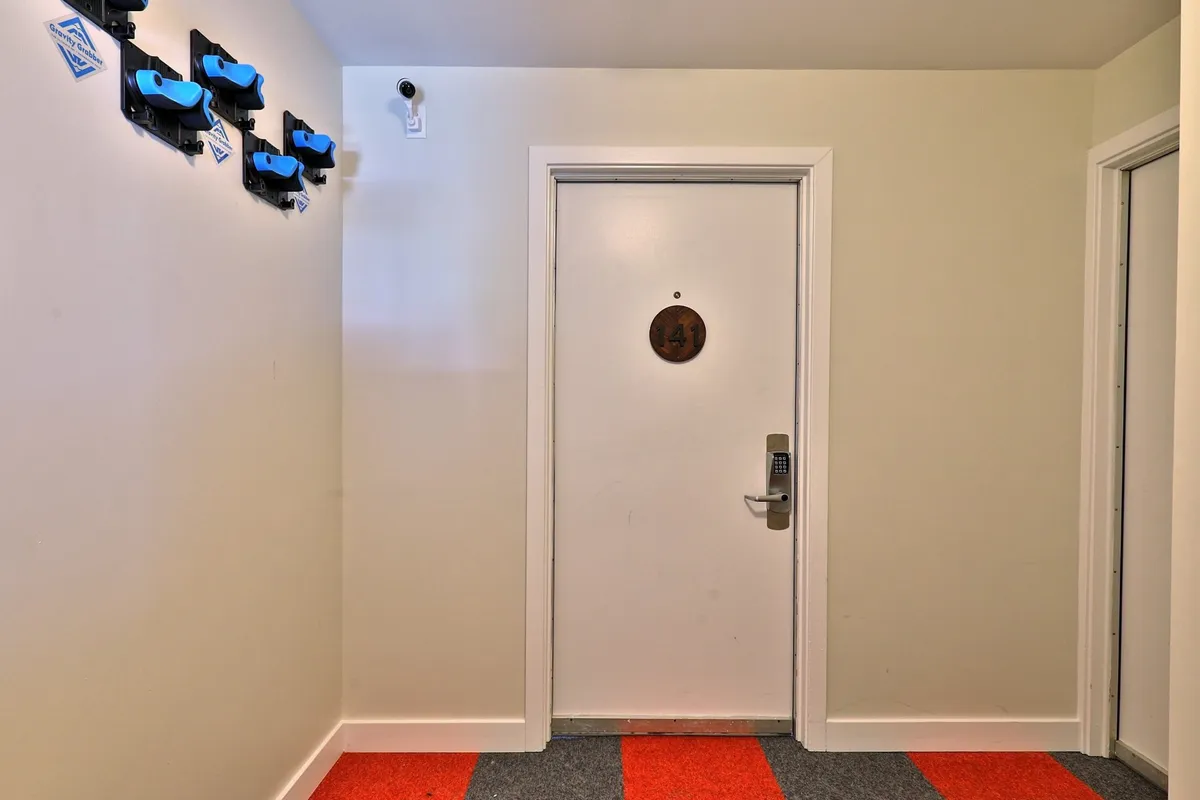 A small entryway with a door bearing a circular number plaque and blue hooks on the wall for hanging items. The red and gray flooring provides a pop of color and an organized feel upon arrival.