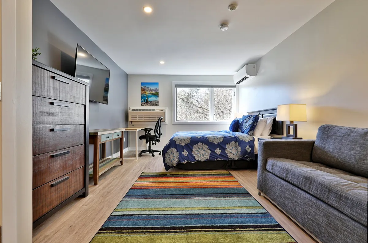 A comfortable bed with floral bedding sits next to a striped area rug, a wall-mounted TV, and a dedicated desk and chair. The workspace is positioned to benefit from natural light, creating a functional yet relaxing corner.