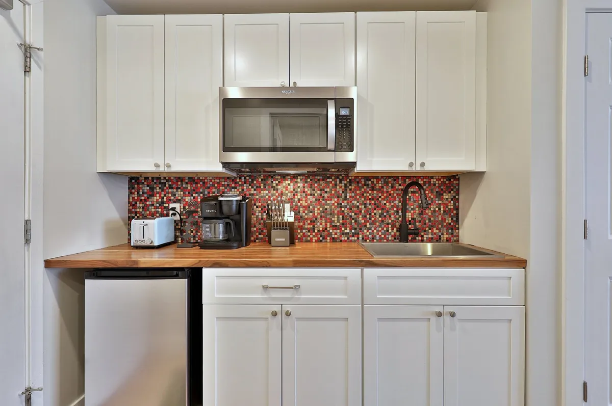 This image features a compact yet inviting kitchenette with white cabinetry, a colorful backsplash, wooden countertop, and visible appliances. The setup showcases a microwave, mini-fridge, sink, and a coffee maker, all neatly integrated into the space. Dishware and utensils are placed within easy reach, highlighting the nook's practical design for quick meal prep.