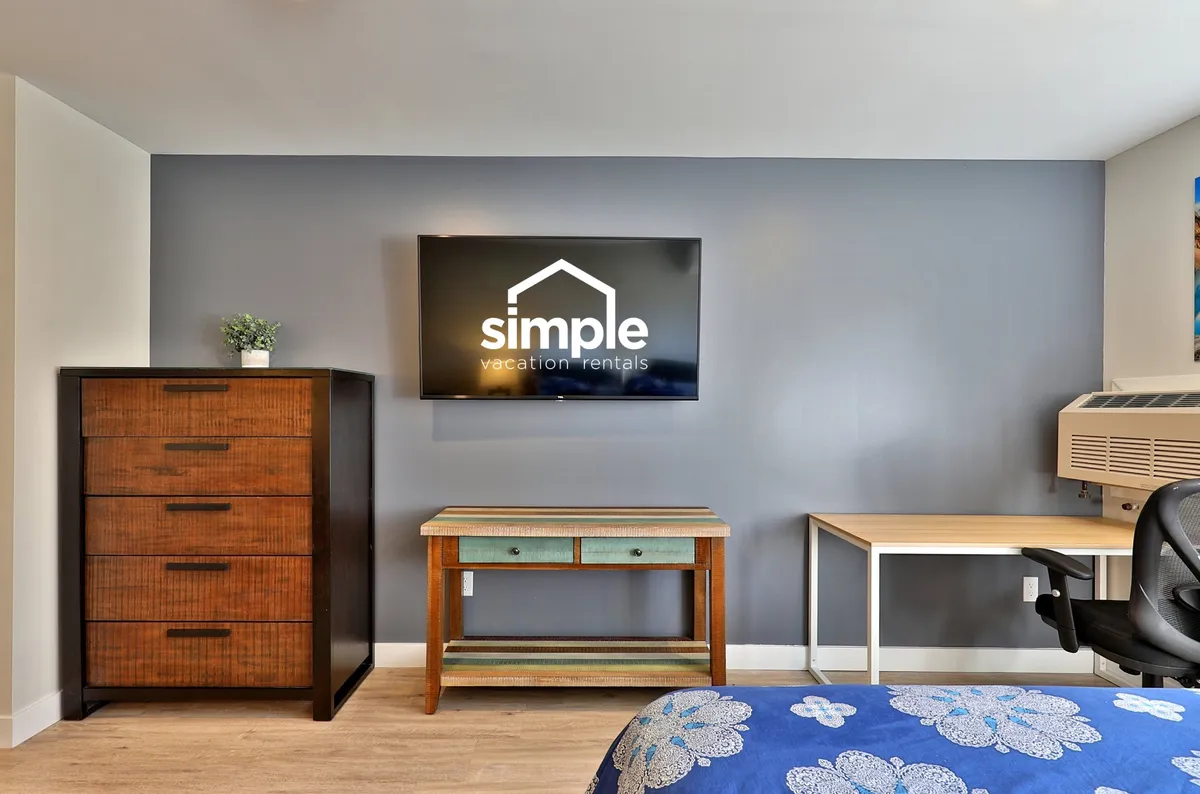A cozy living area featuring a TV with branding, a comfortable chair adjacent to a colorful side table, and a wooden dresser, with the desk area subtly present. The setting suggests a casual space for working, relaxing, and unwinding after a productive day.