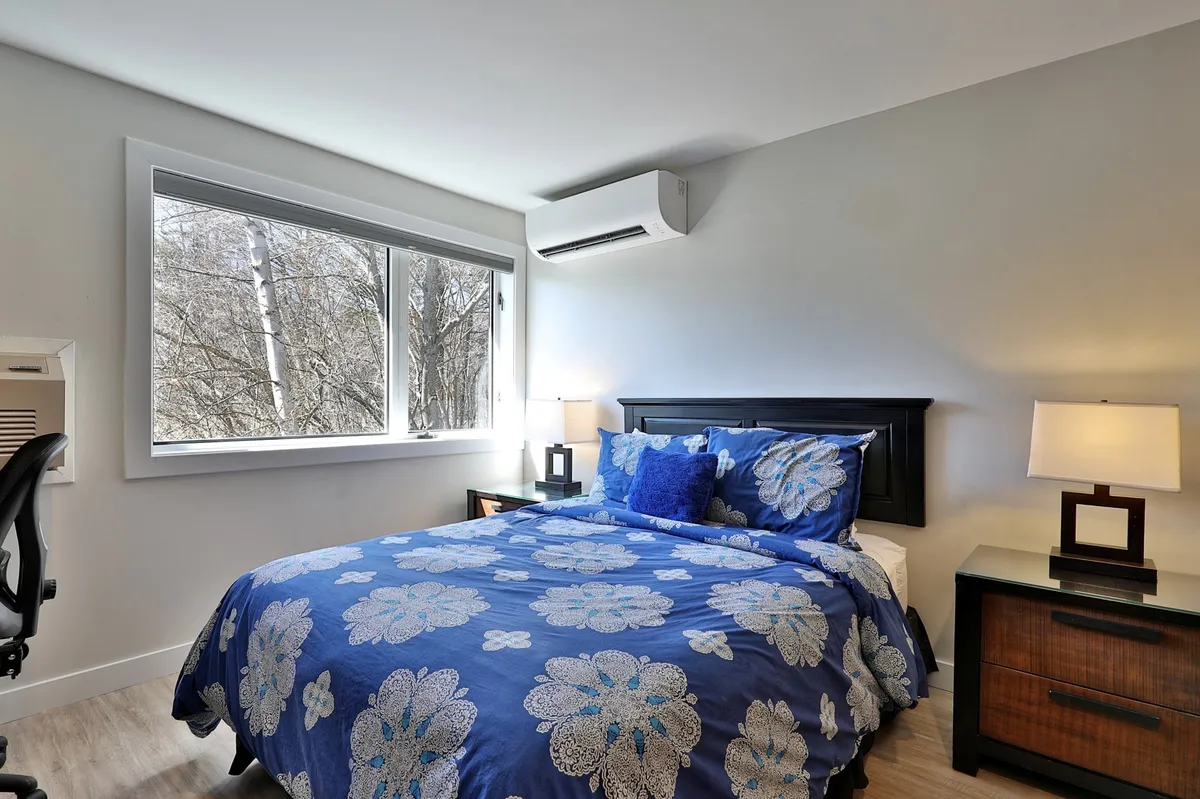 A tranquil bedroom with a bed adorned in blue floral comforter, a desk and chair positioned beside a large window, and a nightstand with a lamp. The window invites sunlight and offers serene garden views, making the workspace both inviting and connected to the outdoors.