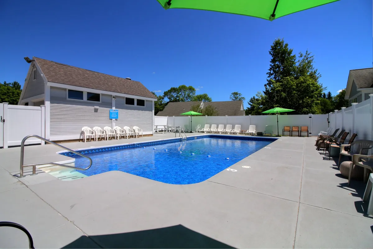 An inviting outdoor swimming pool area surrounded by lush green trees, with lounge chairs and green umbrellas providing a relaxing environment. The space appears tranquil and ideal for leisurely afternoons spent outdoors, either sunbathing, taking a dip, or unwinding with a book.
