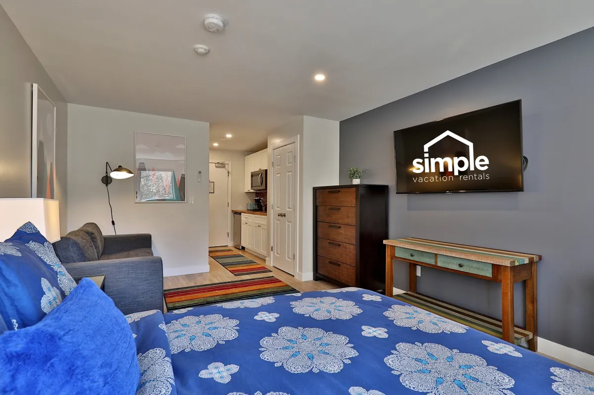 Cozy bedroom with blue floral bedding, a TV displaying a logo, a colorful rug, and a compact dining area. The space feels inviting and flexible—ideal for relaxing after a day of family activities or sharing meals together.