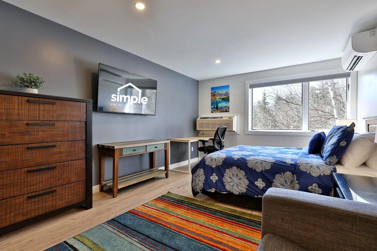 The image shows a cozy studio interior featuring a plush bed with floral blue bedding, a vibrant area rug, a wooden dresser with a TV on top displaying rental branding, and a dedicated desk and chair setup by a window. The light streaming in suggests a welcoming, restful atmosphere ideal for both relaxation and remote work.