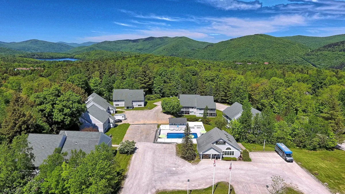 This exterior photo features the main building with a stunning view of the mountains in the background, a swimming pool in the foreground, and lush trees all around. The broad perspective highlights the property's setting within Vermont's natural beauty, reinforcing the location as a hub for year-round recreation and relaxation.