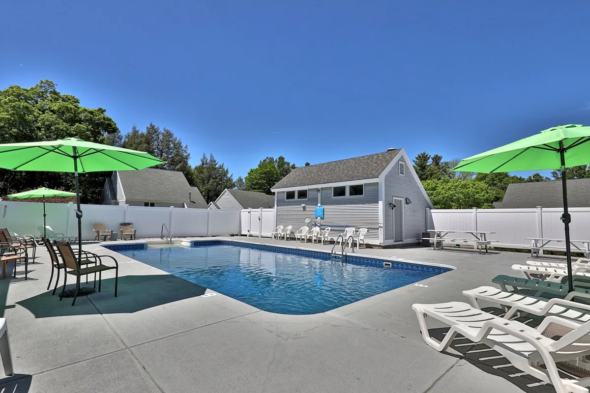 This exterior scene showcases the sparkling swimming pool surrounded by green umbrellas, lounge chairs, and lush trees. Families and friends are seen enjoying themselves by the water, with a changing room building in the background.