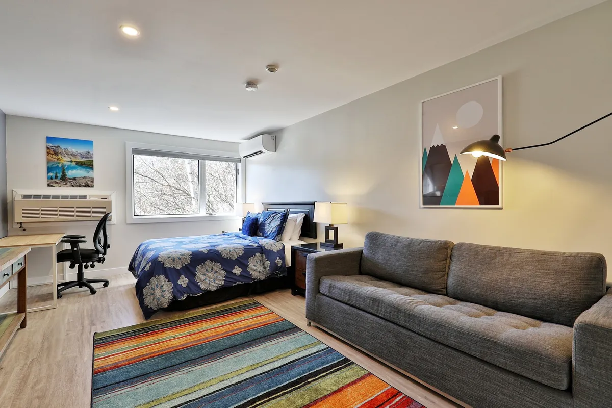 The layout features a bed with floral bedding, a colorful rug, a desk and chair, and cheerful artwork on the wall. The window allows natural light to pour in, making the interior bright and uplifting.