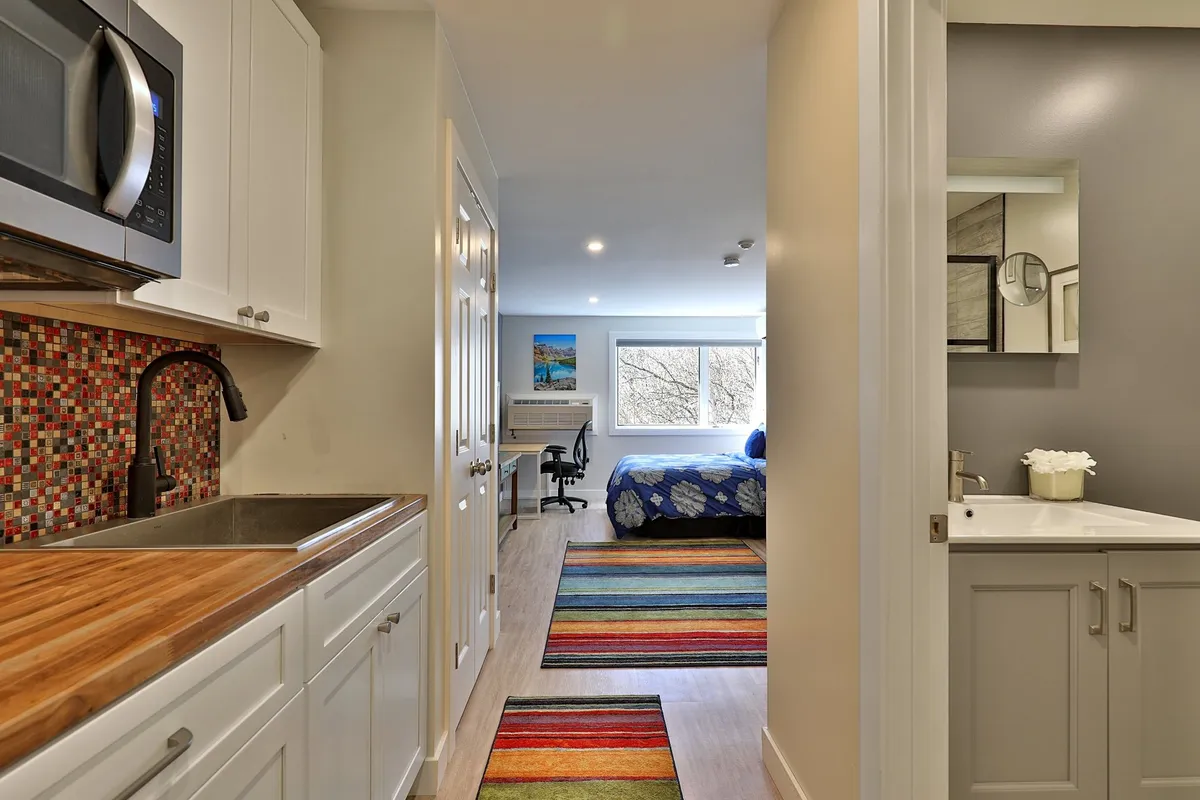 The photo shows the kitchen area integrated into a cozy, vibrant living space. The kitchenette is equipped with essential appliances, and colorful rugs add warmth and personality. The layout makes clear how the kitchenette serves as a central hub for relaxing, cooking, and enjoying daily routines, with the bedroom area and window views nearby.